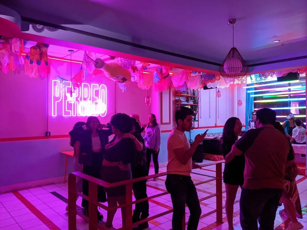Friends dancing under a pink Perreo neon sign and papel picado